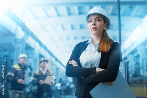 A young successful woman engineer working at a modern plant. In the background two workers. The woman in a white helmet holding technical documentation. Technical revision © ProstoSvet
