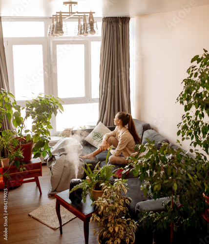 Young woman with long covid syndrome symptoms sitting at couch at home biophilic interior, many indoor trees