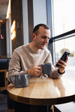 A young man is texting from his mobile phone in a bar and drinking a cappuccino. Young fashionable man drinking espresso coffee in a city cafe during lunch and working