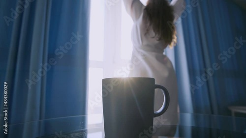 Woman in long bathrobe comes to open blue curtains on large window in spacious hotel room. Lady stretching hands after waking and take mug from glass table, selective focus on cup of coffee