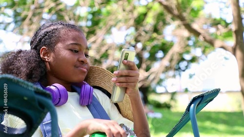 Little Africa American girls sitting on chair using listening favorite songs by headphones while outdoors in a park. Uses communication. learning and playing on the playground