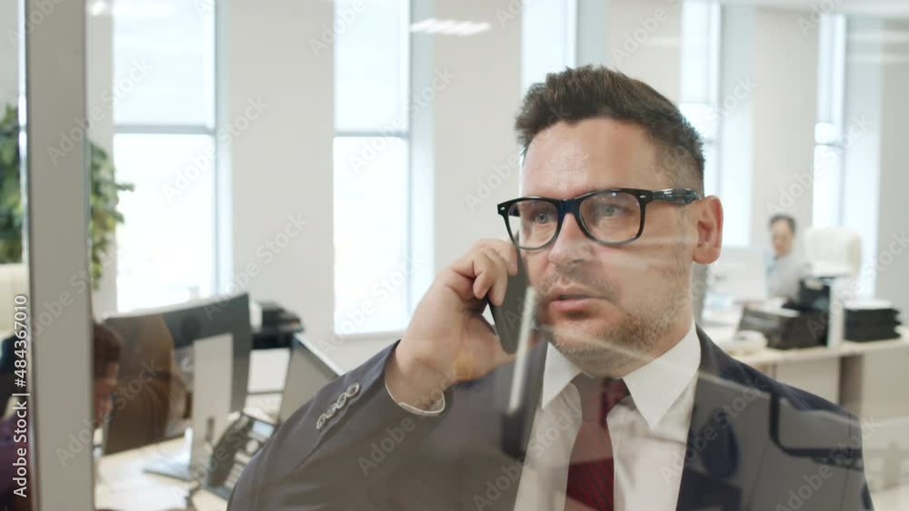 Tracking slowmo close up of male business CEO in suit and glasses standing beside glass wall in office and talking on mobile phone Tracking slowmo close up of male business CEO in suit and glasses standing beside glass wall in office and talking on mobile phone