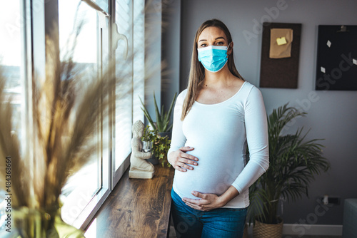 Young beautiful pregnant woman in medical surgical mask and white t-shirt on quarantine at home. Coronavirus, virus, isolation, stay at home. photo