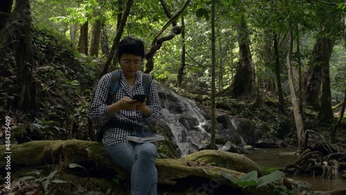 Asian female ecologist sitting on wooden log near waterfall and texting message on smartphone in tropical forest. Woman nature researcher working on mobile phone in the jungle.