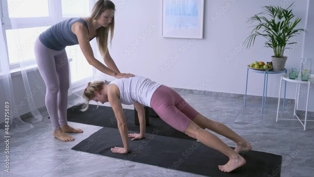 Skilled trainer helps young woman in sportswear to do yoga exercise, assist and supports changing between position at female practice on mat in spacious hall Skilled trainer helps young woman in sportswear to do yoga exercise, assist and supports changing between position at female practice on mat in spacious hall