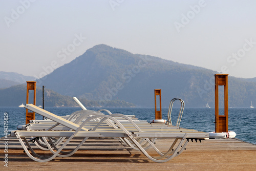 Beach vacation on sea resort, lounge chairs on a wooden pier against mountain. Empty sun beds on green coast background