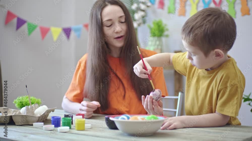 Mom and a son are painting eggs together in a room decorated for the holiday. Concept of family preparation for Easter, festive spring mood Mom and a son are painting eggs together in a room decorated for the holiday. Concept of family preparation for Easter, festive spring mood