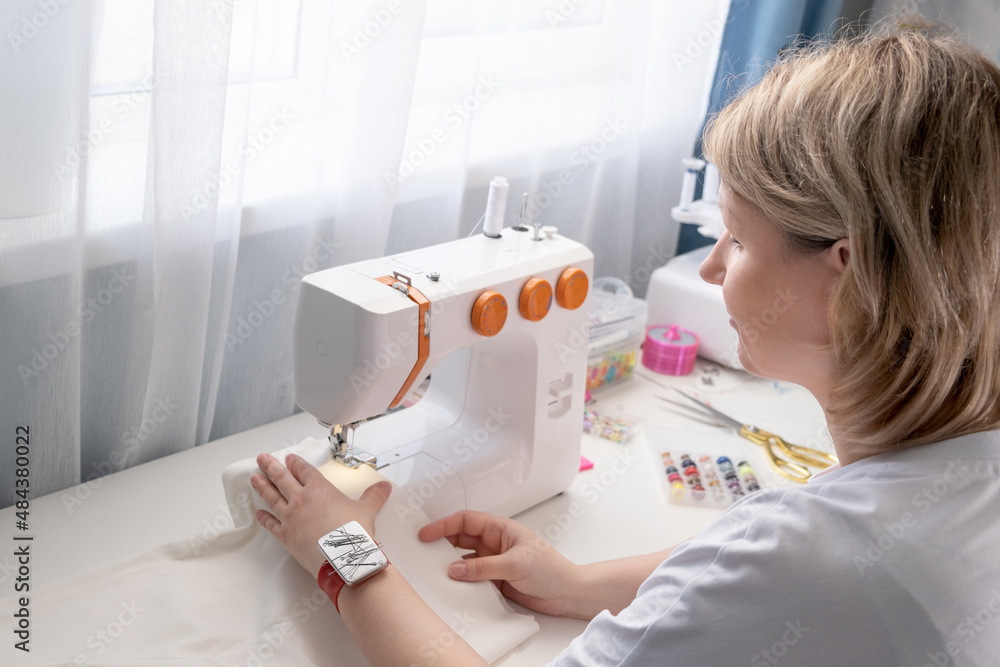 Real middle aged woman tailor sewing clothes using a sewing machine at home. Seamstress working. Hand making clothes in home interior. Female sits enjoying process of sewing clothes. Real middle aged woman tailor sewing clothes using a sewing machine at home. Seamstress working. Hand making clothes in home interior. Female sits enjoying process of sewing clothes.