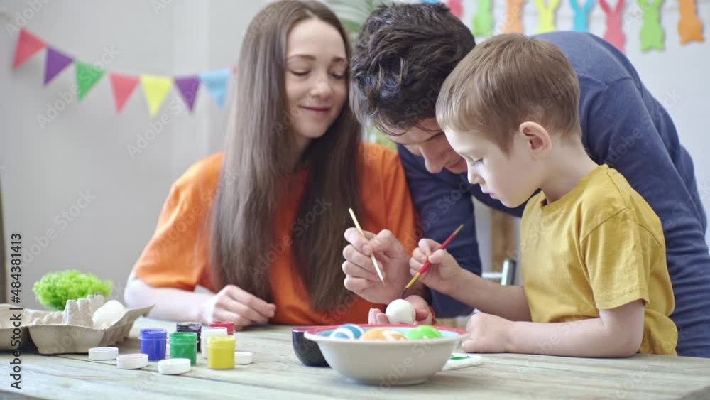 Mom, dad and little son in bright clothes are coloring eggs in a decorated room. Concept of family preparation for Easter, festive spring mood Mom, dad and little son in bright clothes are coloring eggs in a decorated room. Concept of family preparation for Easter, festive spring mood