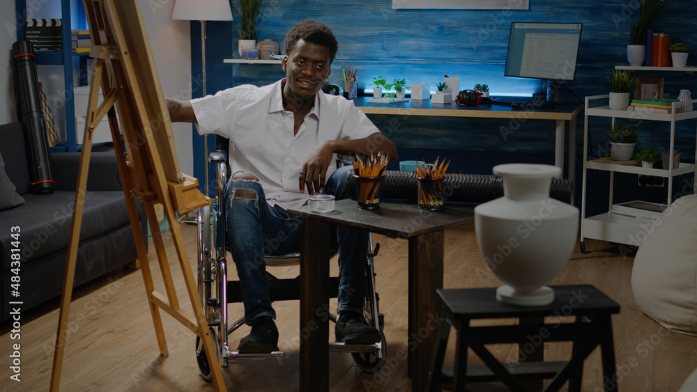 African american man sitting in wheelchair while drawing and designing masterpiece of vase on table. Black young adult using tools, white canvas on easel and colorful pencils African american man sitting in wheelchair while drawing and designing masterpiece of vase on table. Black young adult using tools, white canvas on easel and colorful pencils