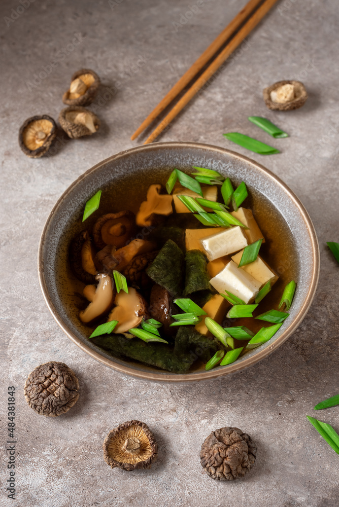 Fototapeta premium Japanese vegan miso soup in a bowl with tofu, shiitake mushrooms, seaweed and green onions. Asian food. Brown grunge background. Side view, selective focus.