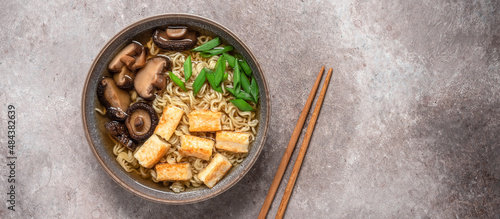 Asian vegan ramen noodle soup with roasted tofu cheese, chives and shiitake mushrooms in a bowl on brown grunge background. Top view. Selective Focus. Banner