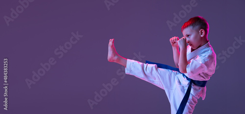 Fotografie Portrait of sportive kid, male taekwondo, karate athletes in doboks doing basic movements isolated on purple background in neon