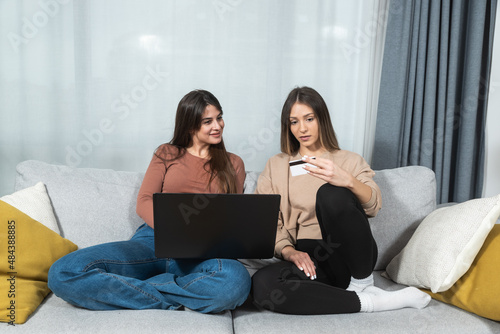 Two happy carefree young women business partners and friends sitting on sofa are happy because they have just tried online shopping with credit card on their new internet store website. 