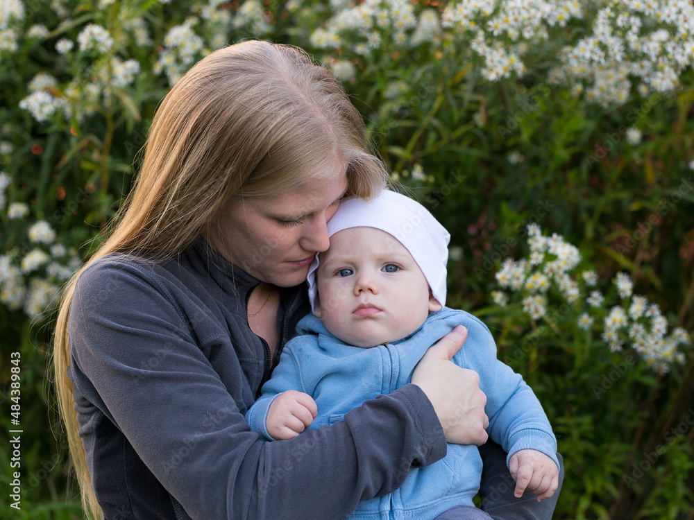 Medium horizontal portrait of pretty young blond mother cuddling baby boy with grumpy expression during late summer afternoon Medium horizontal portrait of pretty young blond mother cuddling baby boy with grumpy expression during late summer afternoon