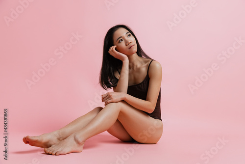 Studio portrait of dreamy looking tender woman sitting and posing in bodysuit isolated over pink background