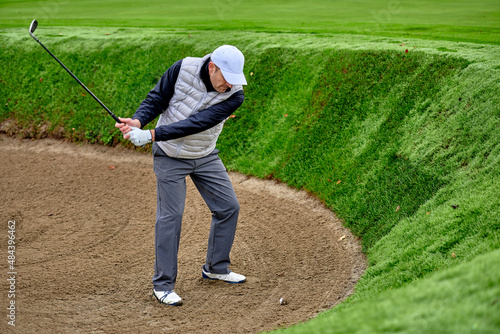The golfer comes out of the bunker by hitting the ball with a golf club, on a winter day with wet grass.