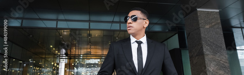 african american security man in formal wear and black sunglasses looking away near hotel entrance, banner.