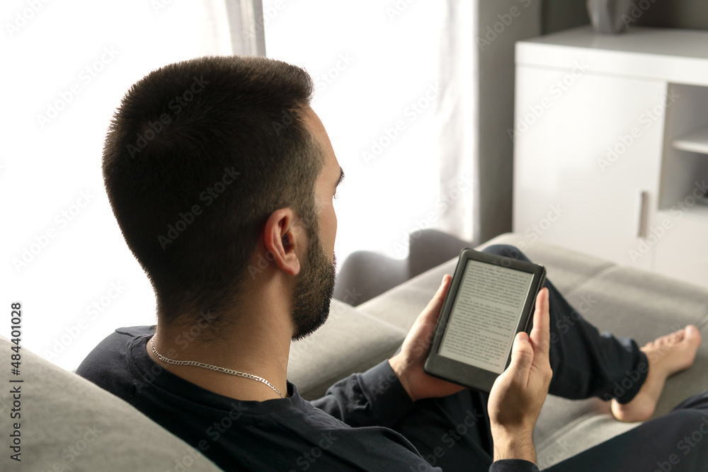 Unrecognizable young man reading with his e-book while sitting on the sofa at home. Unrecognizable young man reading with his e-book while sitting on the sofa at home.