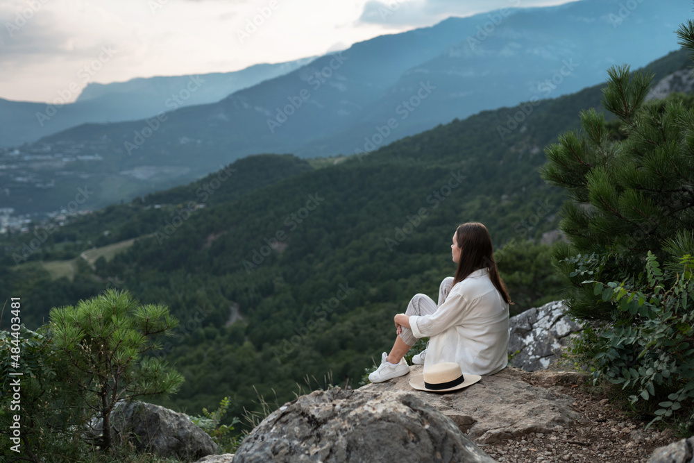 Young beautiful woman with long hair is sitting with her back, meditating and enjoying freedom with a beautiful view of the mountains and the forest. Travel and healthy lifestyle.