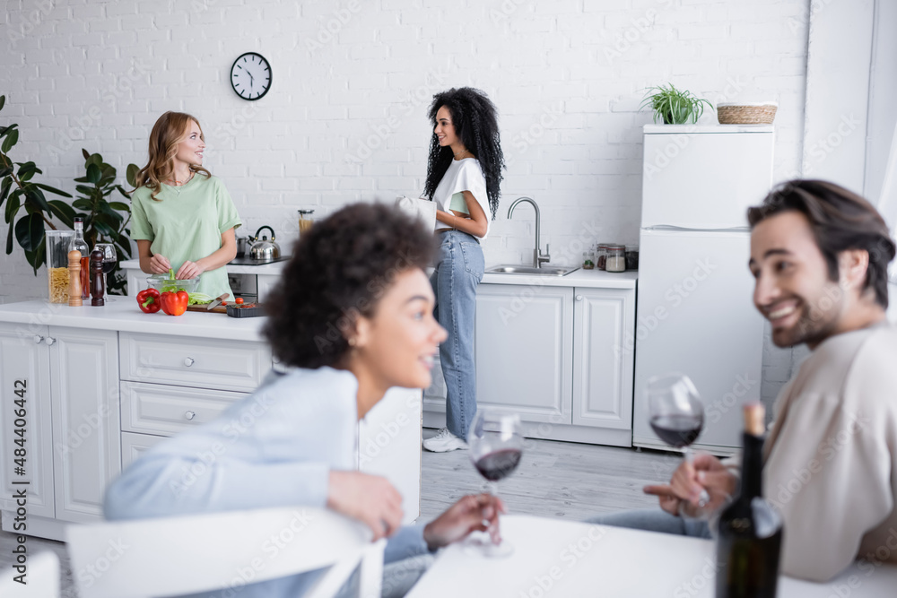 pleased lesbian couple looking at each other near blurred interracial friends with glasses of wine.