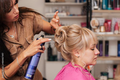 The girl-hairdresser makes the client a hairstyle for a celebration. The hairdresser fixes everything with hairspray