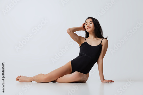 Portrait of young slim asian woman in black bodysuit posing isolated over gray studio background