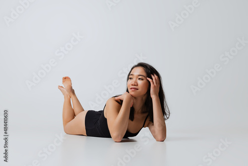 Portrait of beautiful asian woman in black bodysuit lying on floor, posing isolated over gray studio background
