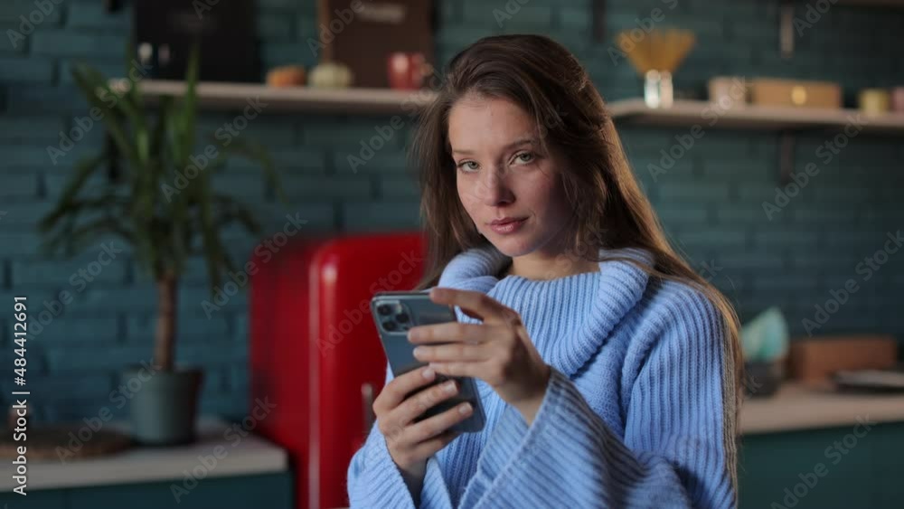 Portrait of young caucasian woman in blue sweater looking at camera while standing indoors with modern smartphone in hands. Concept of people, technology and leisure time. Portrait of young caucasian woman in blue sweater looking at camera while standing indoors with modern smartphone in hands. Concept of people, technology and leisure time.