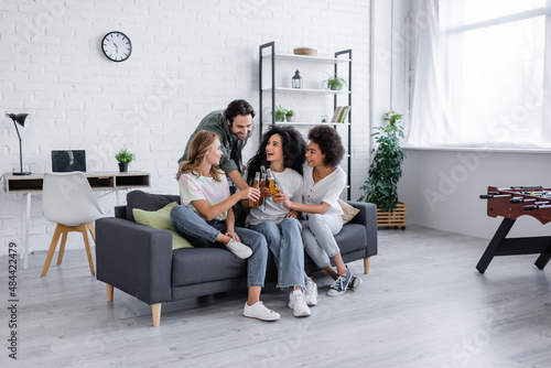 happy man and cheerful interracial women clinking bottles of beer in living room.