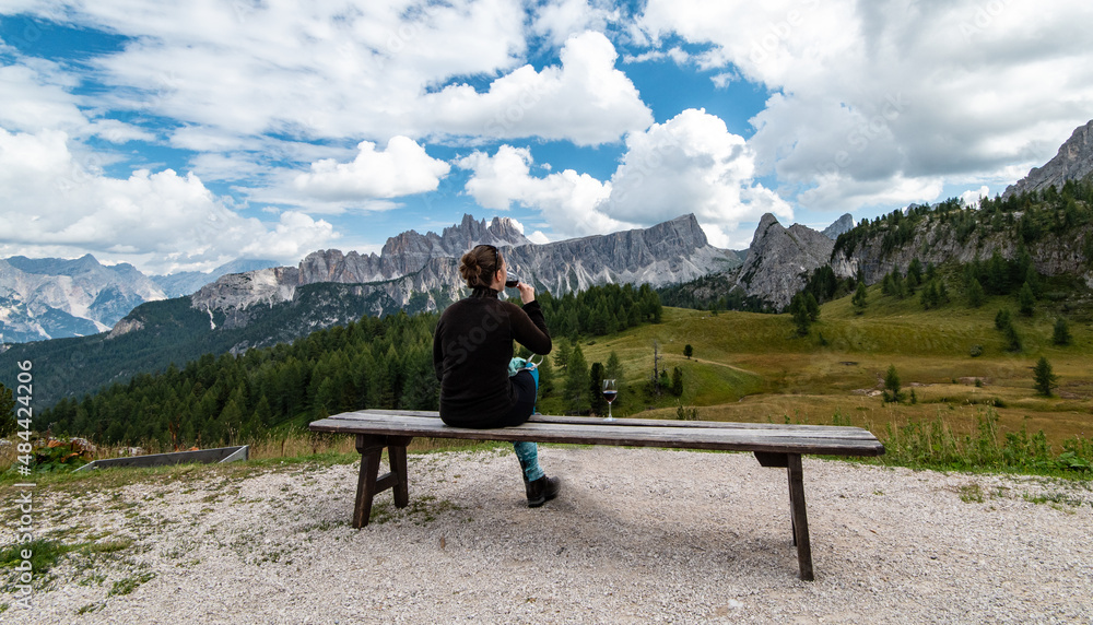 Φωτογραφία Young woman sitting at bench, drinking glass of red wine, looking at Cima Ambrizzola in front of Rifugio Cinque Torri