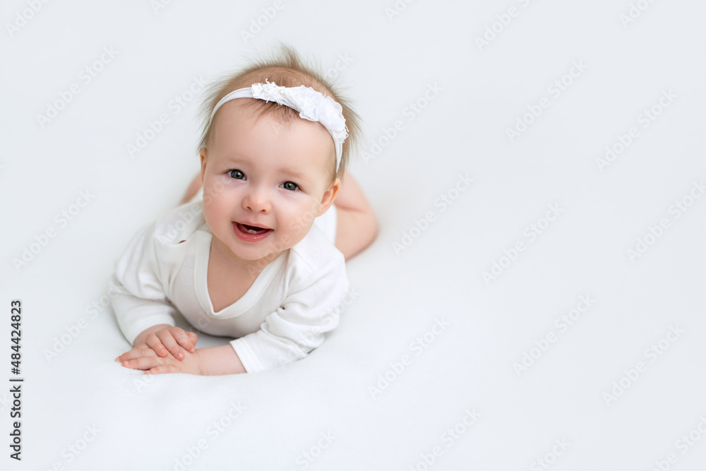 A little six-month-old girl in a white bodysuit and a headband is smiling sweetly lying on her tummy. Happy healthy baby A little six-month-old girl in a white bodysuit and a headband is smiling sweetly lying on her tummy. Happy healthy baby