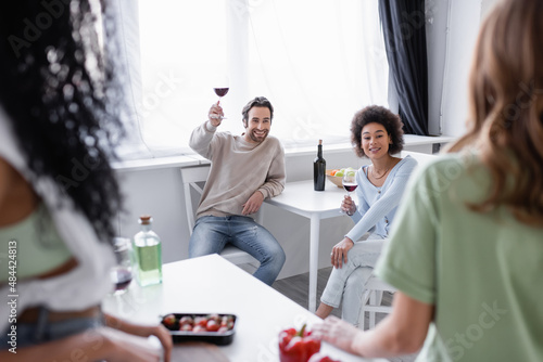 cheerful multiethnic friends holding glasses of red wine near blurred women in modern kitchen.