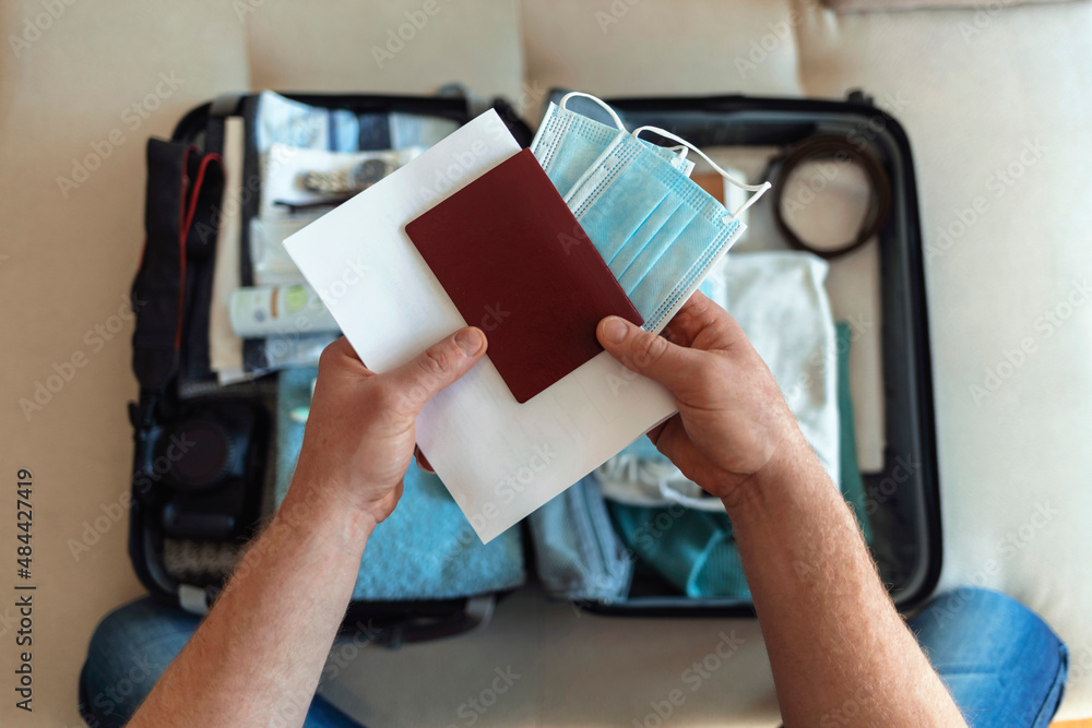 Mens hands putting a face mask and passport in the suitcase prepared to travel, in the new normal, after the coronavirus covid 19 pandemic. High angle view of suitcase with clothes, passport and masks Mens hands putting a face mask and passport in the suitcase prepared to travel, in the new normal, after the coronavirus covid 19 pandemic. High angle view of suitcase with clothes, passport and masks