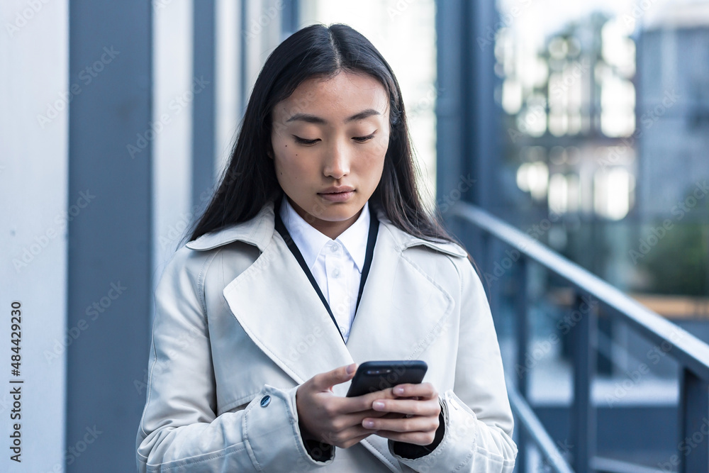 Beautiful Asian woman in business clothes uses the phone, walks near the modern office center outside