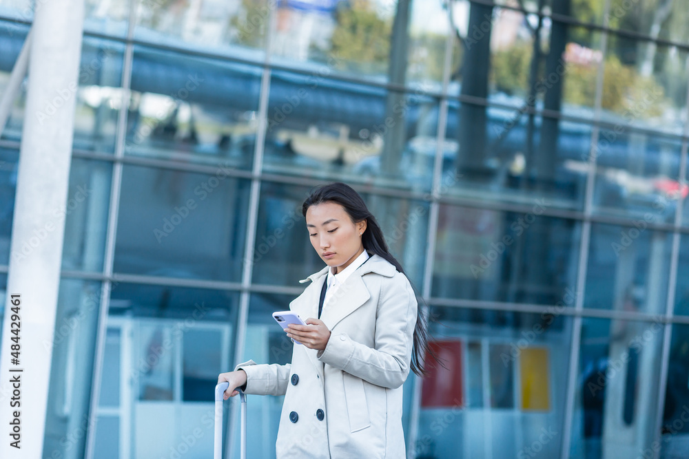 Beautiful Asian woman, near the airport uses a phone application for online booking, a tourist with a large suitcase Beautiful Asian woman, near the airport uses a phone application for online booking, a tourist with a large suitcase