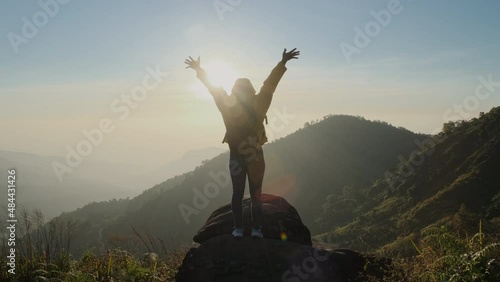 Rear view of a woman in yellow jacket standing on top of mountain summit at sunrise and looking up with raised arms enjoying calm day in the nature breathing fresh air. bio-tourism