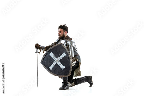 Portrait of serious manm medieval warrior, knight in special protective covering sitting on one knee isolated over white background