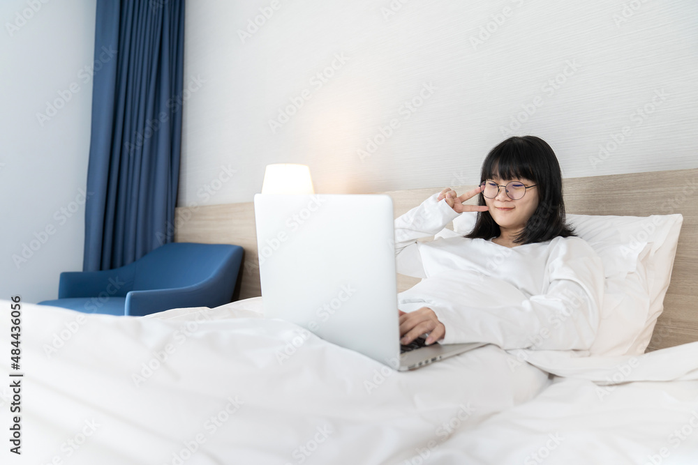 Glasses Asian young pretty teenager rise hand up hi and hello to laptop notebook for meeting and video conference on the bed in Work From Home period.