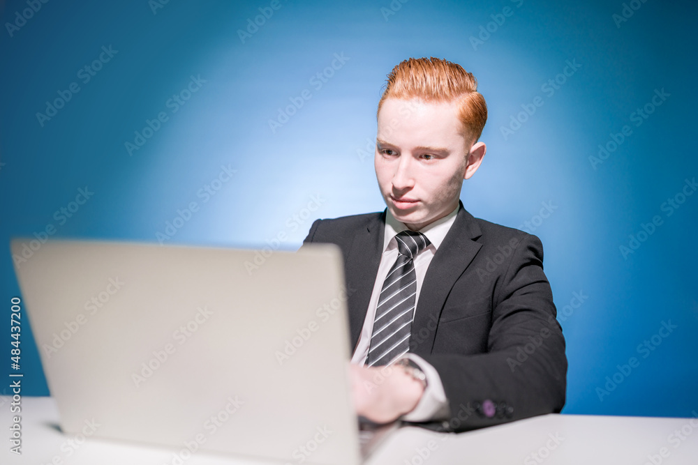 An office worker. The beginning. Seller. Boss. Businessman. Portrait of a young red-haired man in a suit, sitting at a table typing text on a laptop on a blue background. Businessman An office worker. The beginning. Seller. Boss. Businessman. Portrait of a young red-haired man in a suit, sitting at a table typing text on a laptop on a blue background. Businessman