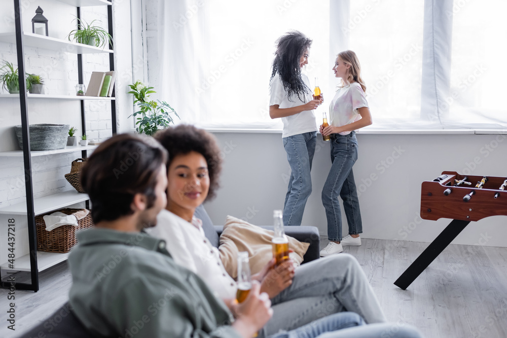 multiethnic lesbian couple holding bottles of beer and standing near window near blurred friends on foreground. multiethnic lesbian couple holding bottles of beer and standing near window near blurred friends on foreground.