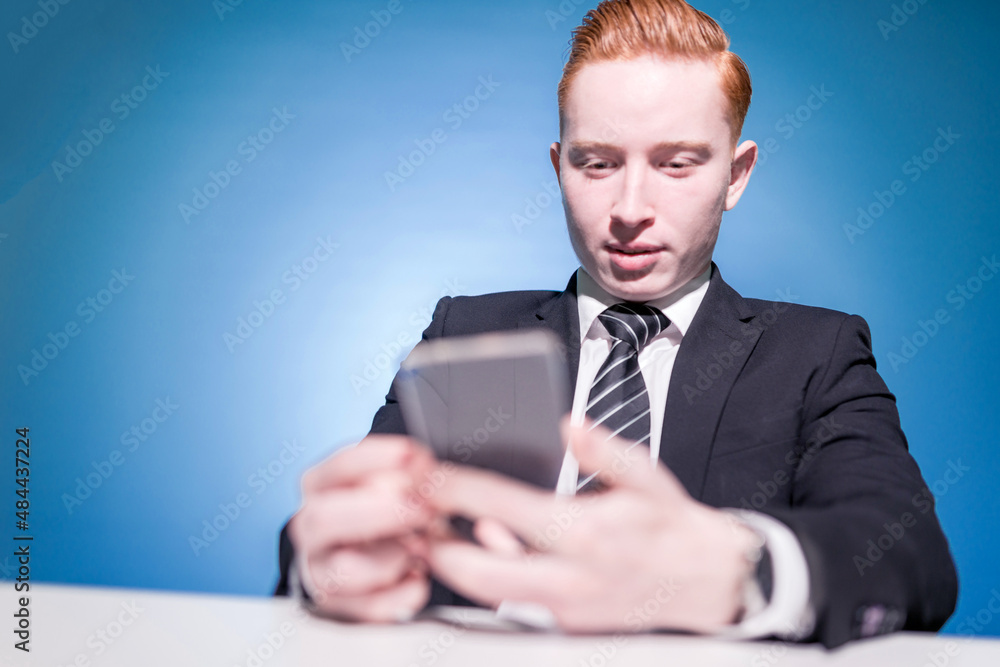 Assistant. Secretary. Portrait of a red-haired man in a suit with a phone in his hands on a blue background, sitting in a typing text. A successful businessman. Seller. Business manager Assistant. Secretary. Portrait of a red-haired man in a suit with a phone in his hands on a blue background, sitting in a typing text. A successful businessman. Seller. Business manager