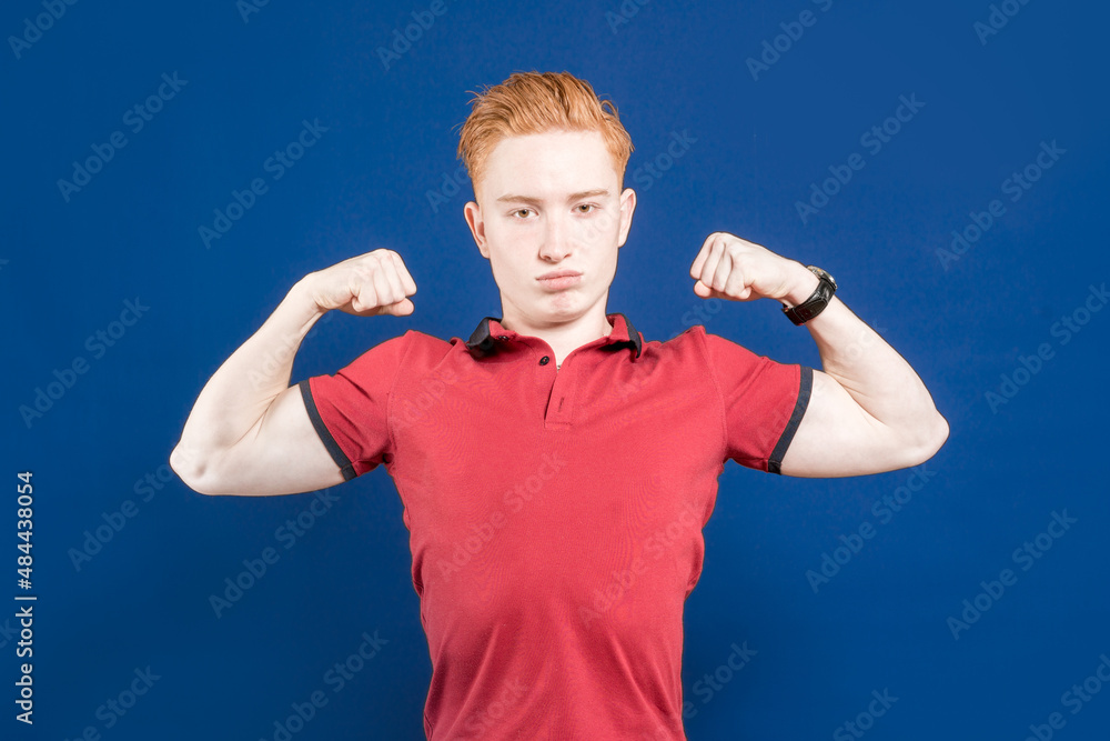 Fototapeta premium A successful smiling male model happily raises her hands. Sportsman. A confident red-haired muscular man, shows muscles on his arms, is proud that he is strong and has strength