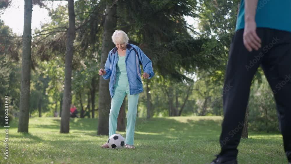 Wide shot portrait of cheerful fit senior woman playing passing soccer ball with unrecognizable man in sunny park. Happy Caucasian wife enjoying sports activity with husband in slow motion Wide shot portrait of cheerful fit senior woman playing passing soccer ball with unrecognizable man in sunny park. Happy Caucasian wife enjoying sports activity with husband in slow motion