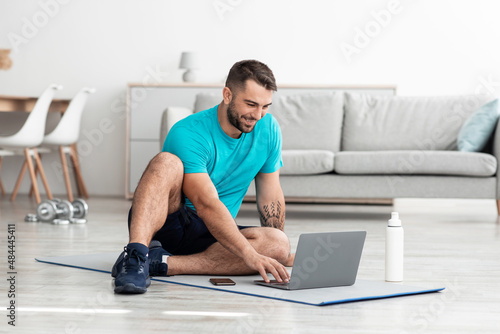 Smiling cheerful millennial muscular caucasian male blogger sitting on mat with bottle of water and computer