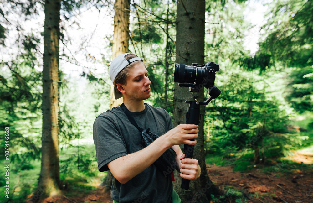 A young male videographer with a serious face stands in a mountain forest with a camera on the stabilizer and adjusts the equipment for shooting. Create travel content for your blog.