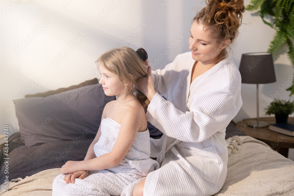 Mother and daughter on the bed in the bedroom. Mom and her little girl are sitting in towels in the room after the bath. Mom takes care of the child, сombing hair with a comb.. Mother and daughter on the bed in the bedroom. Mom and her little girl are sitting in towels in the room after the bath. Mom takes care of the child, сombing hair with a comb..