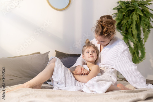 Beautiful mother and cute daughter together. Mom and little girl are sitting on the bed in bath towels in a cozy bedroom on a sunny day..