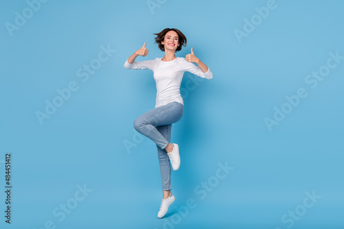 Full size photo of attractive student age girl jumping showing thumbs-up promotion isolated on blue color background