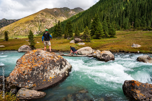 Young man and woman crossing wild turquoise river on tiny wooden bridge  on trail to Ala Kul lake. Karakol valley Issyk-kul region, Ala-kul lake Terskey Alatau mountain range, Kyrgyzstan, Central Asia